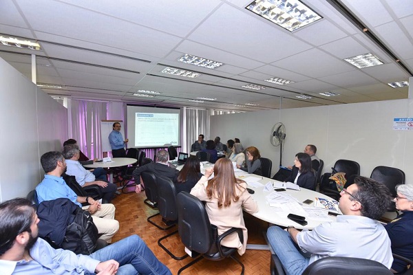 #PraCegoVer: Na foto, um senhor apresenta uma projeção de slide, enquanto outros técnicos e agentes do BID estão sentados em volta da mesa para a reunião
